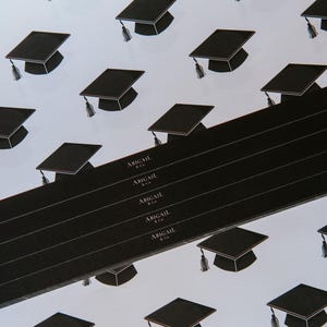 May include: Black and white graduation cap pattern wrapping paper with the text "Abigail & Co." repeated on a black strip.