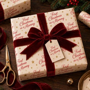 May include: A square gift box wrapped in cream-colored paper with a repeating pattern of gold stars and the red text First Christmas as a Mummy. The box is tied with a deep red velvet ribbon and features a matching gift tag.