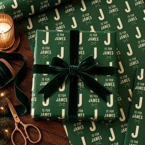 May include: A rectangular gift box wrapped in dark green paper featuring a repeating pattern of the letter J and the text J IS FOR JAMES in white, tied with a dark green velvet ribbon bow centered on the top surface of the package.