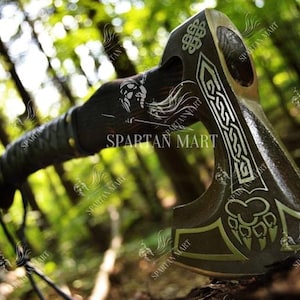 May include: A black and silver axe with a wooden handle. The axe head has a Celtic knot design and a paw print. The axe is resting on a bed of leaves.