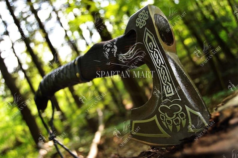 May include: A black and silver axe with a wooden handle. The axe head has a Celtic knot design and a paw print. The axe is resting on a bed of leaves.