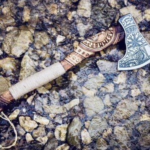 May include: A hand-crafted axe with a wooden handle wrapped in leather. The axe head is decorated with intricate carvings and runes. The axe is lying on a bed of rocks and water.