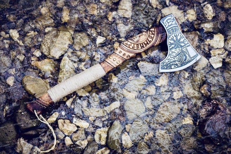May include: A hand-crafted axe with a wooden handle wrapped in leather. The axe head is decorated with intricate carvings and runes. The axe is lying on a bed of rocks and water.