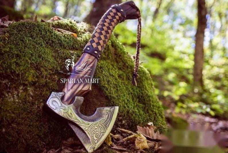 May include: A hand-forged axe with a leather wrapped handle and a decorative metal head. The axe is resting on a mossy rock in a forest setting.