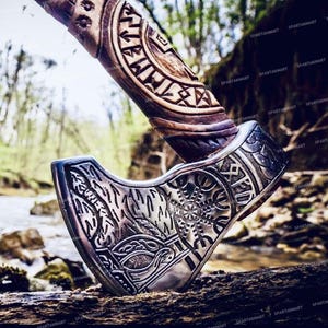 May include: A hand-crafted axe with a wooden handle and a steel head. The handle is carved with intricate designs and the head is decorated with a Viking knotwork pattern. The axe is resting on a log in a forest setting.