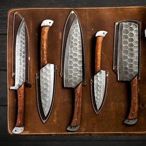 May include: Five kitchen knives with wooden handles are displayed in a brown leather roll. The knives have a hammered finish and are arranged in a row.