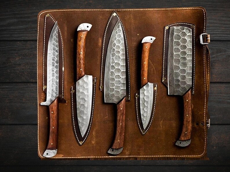 May include: Five kitchen knives with wooden handles are displayed in a brown leather roll. The knives have a hammered finish and are arranged in a row.