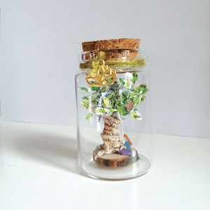 Op de afbeelding: Een glazen pot met een kurken deksel bevat een miniatuur papierboom met groene bladeren en een klein figuurtje dat eronder zit. De pot is versierd met een gouden lotus-hanger en touw.