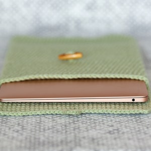 May include: A light green knitted sleeve case with a wooden button closure, holding a rose gold laptop. The case is made of a textured, woven material. The laptop is partially visible, showing its side profile.