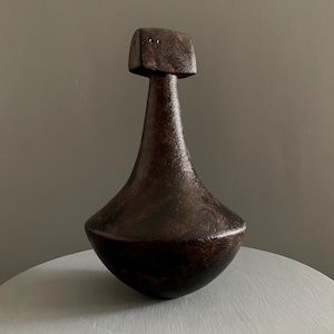 May include: A dark brown decorative object with a distinctive form. It has a rounded base, a long, tapering neck, and a square top. The object appears to be made from a textured material, possibly ceramic or metal, and is likely handmade.