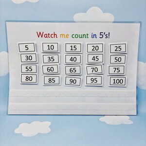 May include: A white board with the text "Watch me count in 5's!" and a grid of numbers from 5 to 100 in increments of 5. The numbers are written in black on white squares.