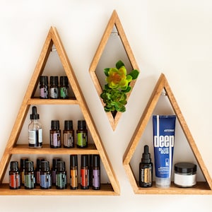 May include: Three wooden triangular shelves with various essential oil bottles and a small potted succulent plant. The shelves are mounted on a white wall. The middle shelf has a clear glass bottle with a dropper. The bottom shelf has a roll-on bottle. The right shelf has a blue tube of "doTERRA deep blue rub" and a small white jar.