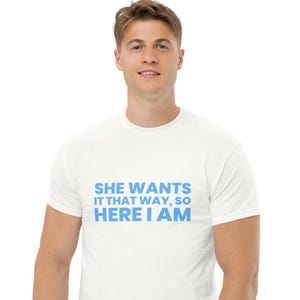 Op de afbeelding: Wit t-shirt met de blauwe tekst: "SHE WANTS IT THAT WAY, SO HERE I AM". Het t-shirt heeft een ronde hals en is waarschijnlijk gemaakt van een zachte stof. De tekst is centraal op de voorkant van het kledingstuk geplaatst.