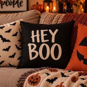 Op de afbeelding: Een vierkant zwart sierkussen met de tekst HEY BOO in een lichtbeige, handgetekend lettertype. Het kussen staat tussen een crèmekleurig kussen met een zwart vleermuispatroon en een oranje kussen met een zwart jack-o'-lantern gezicht.