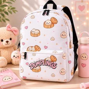 Op de afbeelding: Witte rugzak met een patroon van cartoon dumplings, harten en diamanten. Het woord "DUMPLINGS" is roze gedrukt. De rugzak heeft zwarte ritsen, bandjes en een zijvak. Een dumpling-vormige sleutelhanger is inbegrepen.