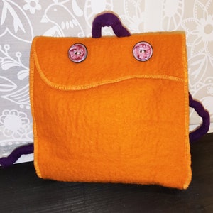Könnte beinhalten: Ein kleiner orangefarbener Filzrucksack mit lila Trägern und zwei rosa Knöpfen.