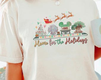 May include: Ivory-colored t-shirt with a colorful graphic featuring a castle, a globe, a Mickey Mouse head, and a tree. The graphic includes Santa in a sleigh and the text "Home for the Holidays". A white mug with a smiley face is also visible.