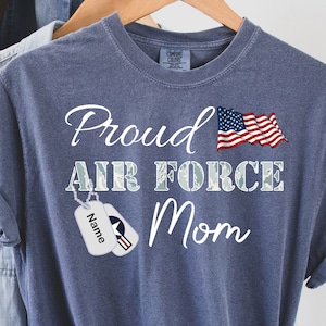 May include: A blue t-shirt with the text "Proud Air Force Mom" and a small American flag. There is a dog tag with the word "Name" on it.