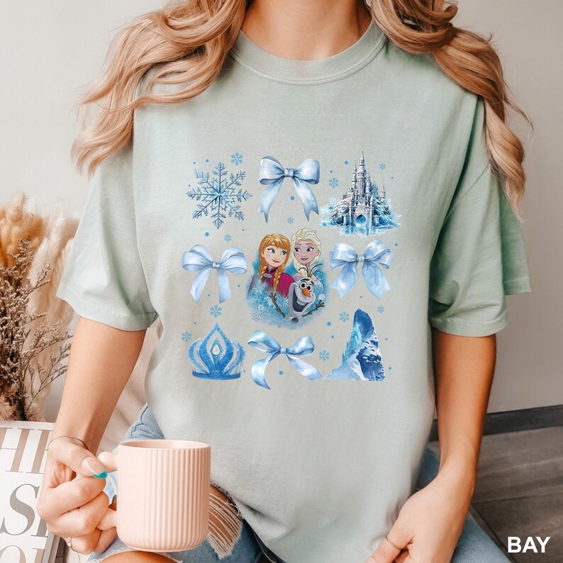 May include: A light green t-shirt featuring a collage of blue Frozen-themed graphics. The design includes images of Anna, Elsa, Olaf, snowflakes, bows, a castle, and a crown. The word "BAY" is printed in white on the bottom right corner.