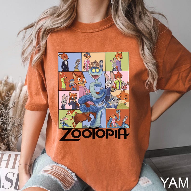 May include: Orange t-shirt featuring a colorful graphic of characters from the animated film "Zootopia." The design includes a grid of images with various animal characters and the film's title in black text. The shirt is a comfortable, casual style.
