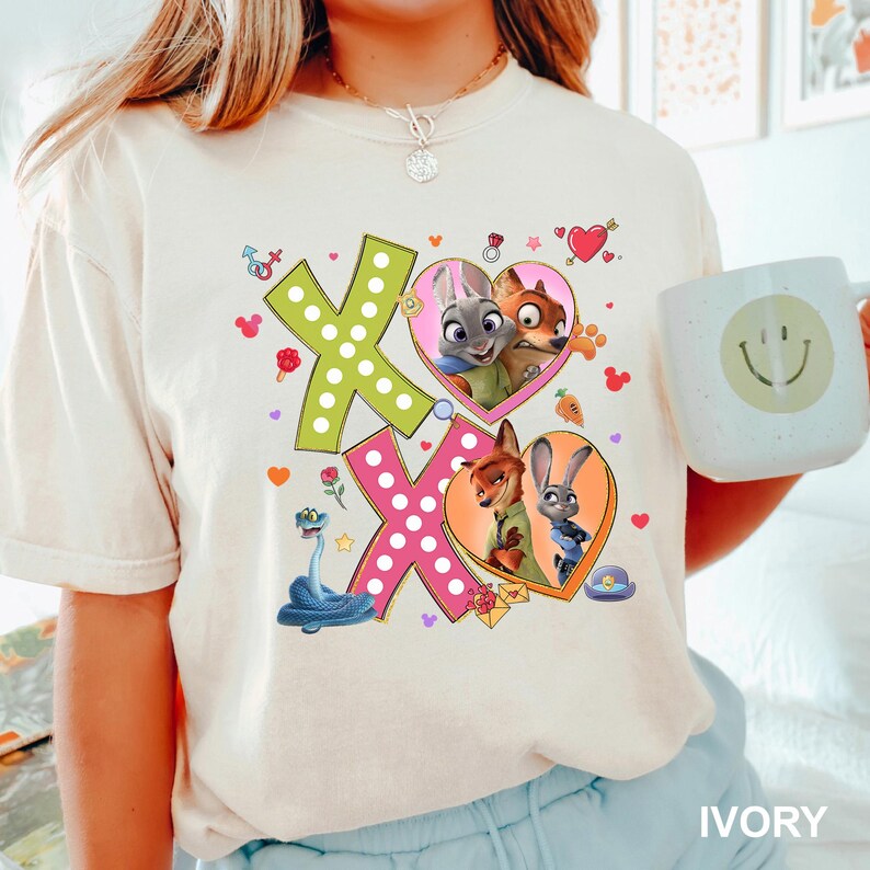 May include: Ivory-colored t-shirt featuring a colorful graphic with the text "XO". The design includes images of characters from the movie Zootopia, hearts, and other decorative elements. The shirt is a comfortable, casual style.
