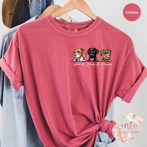 May include: A crimson red t-shirt featuring a graphic of three dogs peeking over a wall. The dogs are a beagle, a black labrador, and a Yorkshire terrier. The text "Arlo & Daisy & Frazier" is printed below the dogs.
