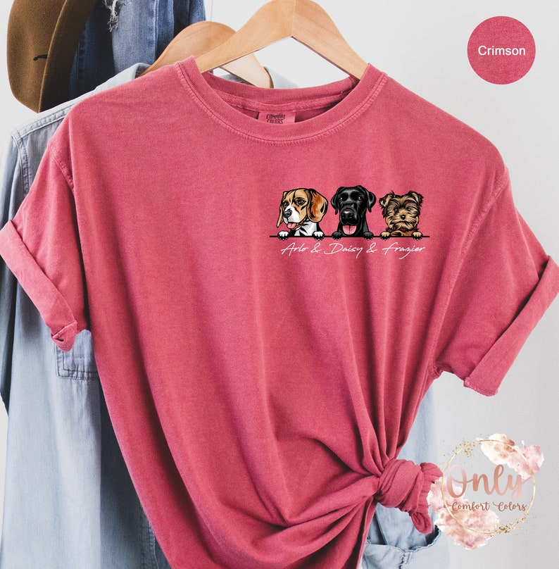 May include: A crimson red t-shirt featuring a graphic of three dogs peeking over a wall. The dogs are a beagle, a black labrador, and a Yorkshire terrier. The text "Arlo & Daisy & Frazier" is printed below the dogs.
