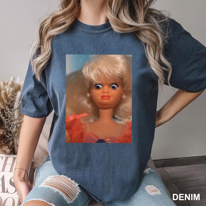 May include: A dark blue short-sleeve t-shirt featuring a rectangular graphic print of a blonde doll with wide, surprised blue eyes and a peach-toned complexion. The doll is wearing a pink garment with ruffled details around the neckline.