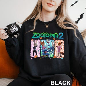 Puede incluir: Sudadera negra con el texto "ZOOTOPIA 2" en verde y naranja. El dise&ntilde;o incluye personajes de dibujos animados en marcos rectangulares coloridos. La palabra "BLACK" est&aacute; impresa en la parte inferior.