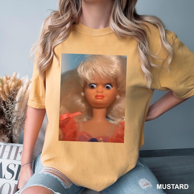 May include: Mustard-colored t-shirt featuring a close-up of a vintage doll with wide eyes and a surprised expression. The doll has blonde hair and is wearing a pink dress. The word "MUSTARD" is printed at the bottom right of the shirt.