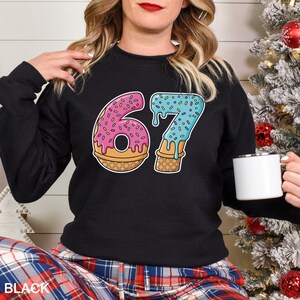 May include: Black sweatshirt with a colorful graphic of the number "67" designed as a donut and ice cream cone. The number "6" is a pink donut with sprinkles, and the "7" is blue ice cream dripping down a cone. The word "BLACK" is printed at the bottom.