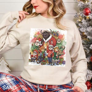 Op de afbeelding: Zandkleurige sweatshirt met een kleurrijke afbeelding van cartoon dieren in feestelijke kleding, waaronder een stier met een kerstmuts, een vos en een konijn. De afbeelding is omgeven door kerstcadeaus en een aquarel achtergrond.