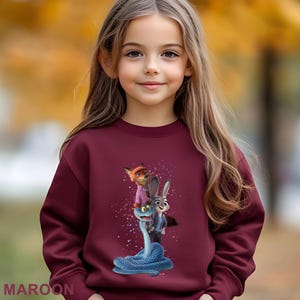 K&ouml;nnte beinhalten: Ein weinrotes Sweatshirt mit einer Grafik von Charakteren aus dem Animationsfilm Zootopia. Die Grafik zeigt Nick Wilde, Judy Hopps und eine blaue Schlange. Das Wort "MAROON" ist unten auf dem Sweatshirt aufgedruckt.
