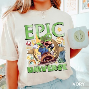 May include: Ivory-colored t-shirt featuring a green and brown graphic of a cartoon character with the words "EPIC UNIVERSE" in green. A white mug with a smiley face is held in the image.