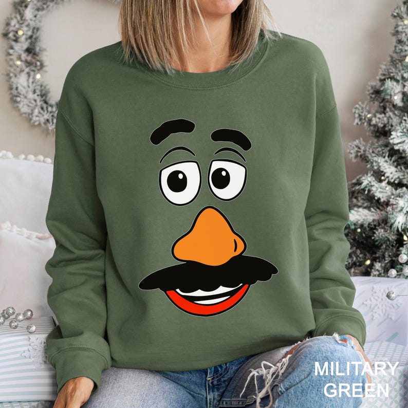 May include: Military green sweatshirt featuring a cartoon face with a large orange nose, black mustache, and a red smile. The eyes are white with black pupils and black eyebrows. The text "MILITARY GREEN" is at the bottom right.