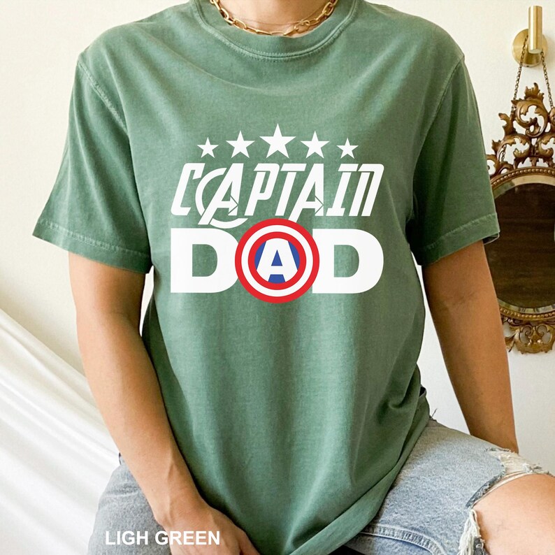 Puede incluir: Camiseta verde claro con el texto blanco "CAPTAIN DAD", con un emblema de escudo rojo, blanco y azul. Encima del texto hay cinco estrellas blancas. La camiseta es de estilo c&oacute;modo e informal.