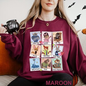Puede incluir: Sudadera granate con una cuadr&iacute;cula de retratos de personajes de dibujos animados, incluyendo Chief Bogo, Nick Wilde y Judy Hopps. La palabra "MAROON" est&aacute; impresa en la parte inferior. La persona sostiene una taza negra que dice "Witches Brew".