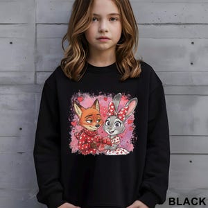 K&ouml;nnte beinhalten: Schwarzes Sweatshirt mit einem Cartoon-Fuchs und -Hasen, die ein herzf&ouml;rmiges Geschenk teilen, umgeben von Herzen. Der Fuchs tr&auml;gt einen roten Schal, und der Hase hat eine rote Schleife. Das Wort "BLACK" ist unten rechts in Wei&szlig; aufgedruckt.