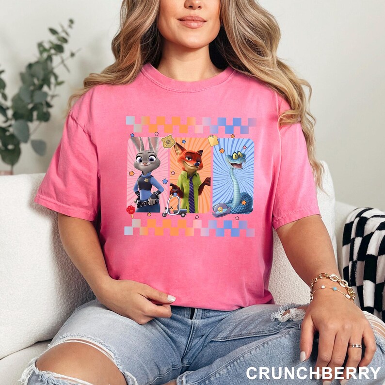 May include: A bright pink short-sleeved t-shirt featuring a graphic print of three animated characters from Zootopia: Judy Hopps, Nick Wilde, and a blue snake. The design is framed by a colorful checkered border and includes small star icons.