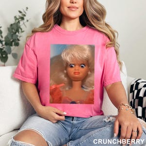 May include: A pink t-shirt featuring a framed image of a vintage doll with wide eyes and blonde hair. The text "CRUNCHBERRY" is printed in white at the bottom right corner of the image.