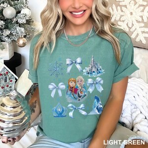 May include: A light green t-shirt featuring a graphic of characters and elements from the movie Frozen. The design includes snowflakes, bows, a castle, and the text "LIGHT GREEN".