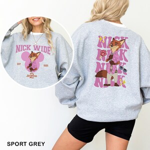 K&ouml;nnte beinhalten: Sportgraues Sweatshirt mit einem Cartoon-Fuchs-Charakter in einem rosa Hemd und Sonnenbrille. Das Design auf der Vorderseite zeigt den Text "NICK WIDE" in Rosa, mit "EST 2025" und "Zootopia 2" darunter. Auf der R&uuml;ckseite befindet sich ein gr&ouml;&szlig;eres Bild des Fuchses und der Name "NICK".