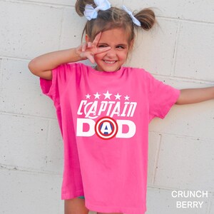 Puede incluir: Una camiseta rosa brillante con las palabras "CAPTAIN DAD" en blanco, con un gr&aacute;fico de escudo con la letra "A" en el centro. La camiseta la lleva una persona joven con el pelo claro peinado en dos mo&ntilde;os y lazos blancos. El color de la camiseta es "Crunch Berry".