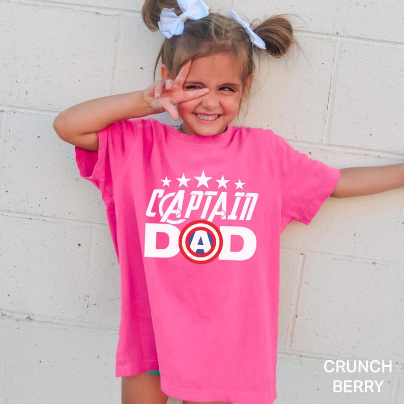 Puede incluir: Una camiseta rosa brillante con las palabras "CAPTAIN DAD" en blanco, con un gr&aacute;fico de escudo con la letra "A" en el centro. La camiseta la lleva una persona joven con el pelo claro peinado en dos mo&ntilde;os y lazos blancos. El color de la camiseta es "Crunch Berry".
