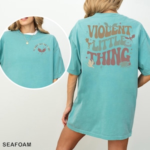 May include: Seafoam green t-shirt with the text "VIOLENT LITTLE THING" on the back in brown, green, and pink. The front of the shirt has the text "VI BES LIT GET LIT LIKE" and a dragon graphic. The shirt is a relaxed fit.