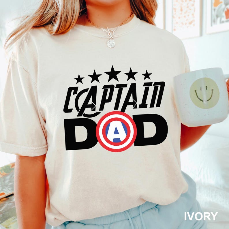 Puede incluir: Camiseta color marfil con el texto "CAPTAIN DAD" en negro, con un dise&ntilde;o de escudo rojo, blanco y azul. Encima del texto hay cinco estrellas negras. En la imagen se sostiene una taza blanca con una carita sonriente.