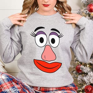 May include: Sport Grey sweatshirt featuring a cartoon face with large eyes, a pink nose, and a red mouth. The sweatshirt is a light gray color. The text "Sport Grey" is at the bottom of the image.