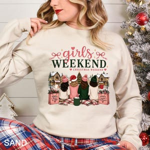 May include: A sand-colored crewneck sweatshirt featuring a graphic print of four people sitting together. The text reads "girls WEEKEND" and "CHRISTMAS WEEKEND" above an illustration of a snowy village scene with trees, houses, and shopping bags.