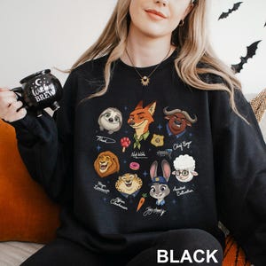 May include: Black sweatshirt featuring cartoon illustrations of characters from a popular animated movie. The design includes a sloth, fox, lion, and other animal characters. The word "BLACK" is printed at the bottom of the sweatshirt.