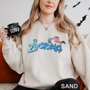Puede incluir: Sudadera de color arena con la palabra "Zootopia" en azul, con personajes de dibujos animados. La sudadera tiene mangas largas y cuello redondo. La palabra "SAND" est&aacute; impresa en la parte inferior de la imagen.